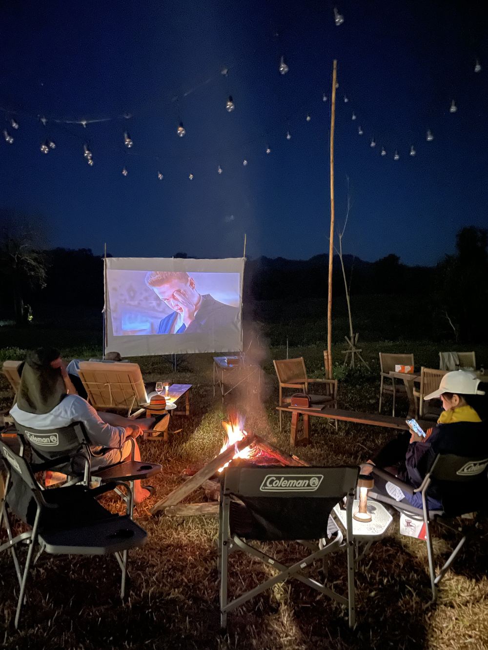 Guests around the bonfire watching an outdoor film under string lights